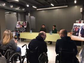 Pokaż zdjęcie: Zdjęcie wykonane z widowni. Na pierwszym planie siedzą trzy osoby niepełnosprawne na wózkach, dwie z nich pochylają się do siebie rozmawiając. W tle długi stół z którym siedzą ludzie. Obok niego siedzi osoba na wózku inwalidzkim elektrycznym.