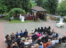 Pokaż zdjęcie: Zdjęcie z lotu ptaka przedstawiające uroczystość jubileuszu 20-lecia Warsztatu Terapii Zajęciowej w Siemiatyczach. Na scenie, pod zieloną altaną, przemawia kobieta w jasnym stroju obok siostry zakonnej i grupy uczestników warsztatu w czarno-czerwonych strojach. Przed sceną siedzi kilkadziesiąt osób, w tym siostry zakonne i zaproszeni goście.