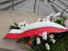 Pokaż zdjęcie: Przed pomnikiem leży wiązanka kwiatów w kolorach: białym i czerwonym. Na niej znajduje się szarfa z napisem: "Państwowy Fundusz Rehabilitacji Osób Niepełnosprawnych". 