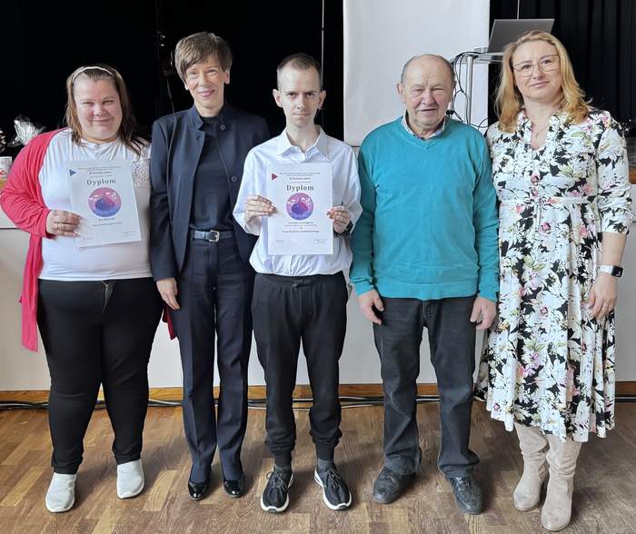 Pokaż zdjęcie: Pięć osób stoi w rzędzie, prezentując dyplomy. Od lewej: kobieta w białej bluzce, różowym swetrze i czarnych spodniach trzyma dyplom. Obok niej kobieta w czarnej marynarce i spodniach. Następnie mężczyzna w jasnoniebieskiej koszuli i sportowych butach, również trzymający dyplom. Dalej starszy mężczyzna w turkusowym swetrze oraz kobieta o blond włosach w jasnej sukience w kwiaty i okularach.