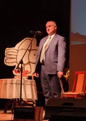 Pokaż zdjęcie: Paweł Wdówik, wiceminister, pełnomocnik rządu ds. osób niepełnosprawnych. Pełnomocnik stoi na scenie, towarzyszy mu pies przewodnik.