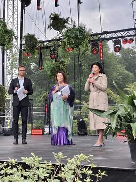 Scena w ogrodzie botanicznym otoczona kwiatami i roślinami. Na scenie mężczyzna i dwie kobiety przemawiają do mikrofonów.