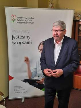 Pokaż zdjęcie: Na tle plakatu stoi mężczyzna w garniturze. Na bilbordzie jest pani na wózku inwalidzkim i inrormacje dotyczące PFRON. 