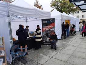 Pokaż zdjęcie: Na dziedzińcu stoją ustawione w rzędzie białe namioty. Pod nimi są stoiska różnych instytucji i placówek. Przed namiotami znajdują się ludzie. 