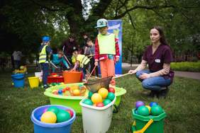 Pokaż zdjęcie: Kobieta kuca i trzyma we ręku siatkę na kiju, obok stoi chłopiec w czapce z daszkiem. Przed nimi basenik z wodą i kulki w wiaderkach