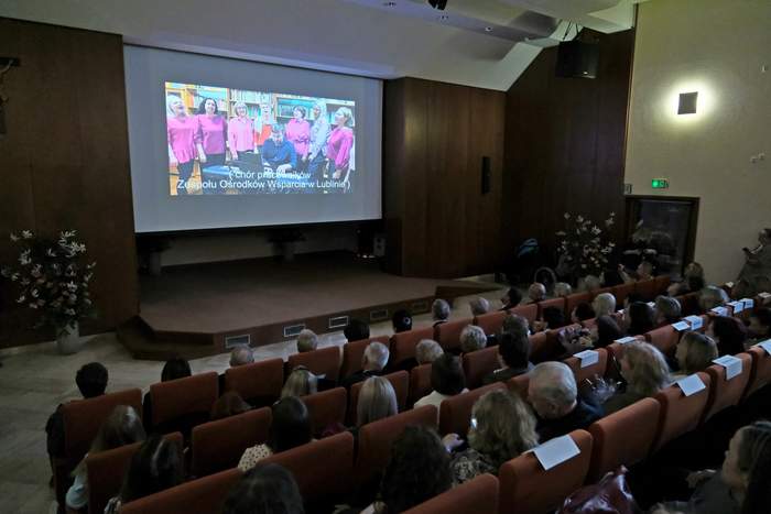 Pokaż zdjęcie: Widok sali widowiskowej wypełnionej publicznością. Na scenie ekran z projekcją występu chóru pracowników Zespołu Ośrodków Wsparcia w Lublinie. Uczestnicy siedzą w rzędach foteli.