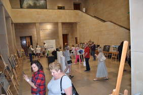 Pokaż zdjęcie: Przestrzeń muzeum z ekspozycją prac konkursowych, ustawionych na sztalugach i kubikach. Grupa zwiedzających. W tle banner PFRON