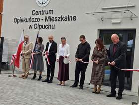 Grupa siedmiu osób stoi przed wejściem do Centrum Opiekuńczo-Mieszkalnego w Olpuchu i wspólnie przecina biało-czerwoną wstęgę. Obok znajduje się flaga Polski.