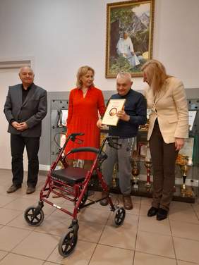 Pokaż zdjęcie: Na zdjęciu 4 osoby, dwóch mężczyzn i dwie kobiety. Mężczyźnie przed którym stoi chodzik wręczono dyplom, który trzyma w dłoniach i prezentuje