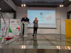 Pokaż zdjęcie: Na podwyższonej szarej scenie, przy mównicy stoi kobieta i mężczyzna. 