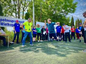 Pokaż zdjęcie: Mężczyzna skacząc w dal unosi się nad ziemią. Zawodnikowi kibicują osoby zgromadzone na boisku wyłożonym sztuczną trawą. W tle wysokie metalowe ogrodzenie, a na nim zawieszone są banery promujące imprezę sportową. Za płotem rosną wysokie drzewa.