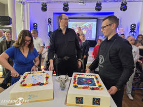 Pokaż zdjęcie: Dwóch mężczyzn oraz jedna kobieta przymierzają się do pokrojenia na mniejsze kawałki dwóch tortów okolicznościowych z okazji jubileuszu 20-lecia Środowiskowego Domu Samopomocy we Włoszakowicach. W tle znajdują się goście zaproszeni na uroczystość.