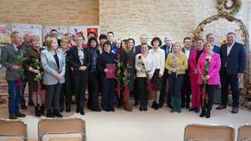 Pokaż zdjęcie: Na środku sali stoją w dwóch rzędach ludzie. Są elegancko ubrani. Niektóre osoby trzymają w dłoniach róże. 