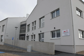 Nowoczesny, jasny budynek z dwoma kondygnacjami, dużymi oknami i łącznikiem przeszklonym; przed nim chodnik, znak parkingu i niski betonowy mur, na ścianie tablica z godłem Polski.  