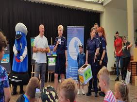 Pokaż zdjęcie: scena na której stoją przedstawiciele policji i powiatu oraz dzieci które otrzymały nagrodę, za uczestnictwo w konkursie, po lewej stronie stoi maskotka przebrana za policjanta  