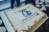 Duży prostokątny tort z jasnym kremem, udekorowany na brzegach owocami i rozetkami. Na środku znajduje się niebieski napis „75 lat” oraz okrągłe logo z podpisem „OKRĘG WIELKOPOLSKI”. W tle widać talerzyki z pokrojonymi kawałkami ciasta oraz stojące obok osoby.