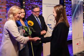 Pokaż zdjęcie: Na scenie odbywa się wręczenie nagród, dwie kobiety podają sobie ręce.