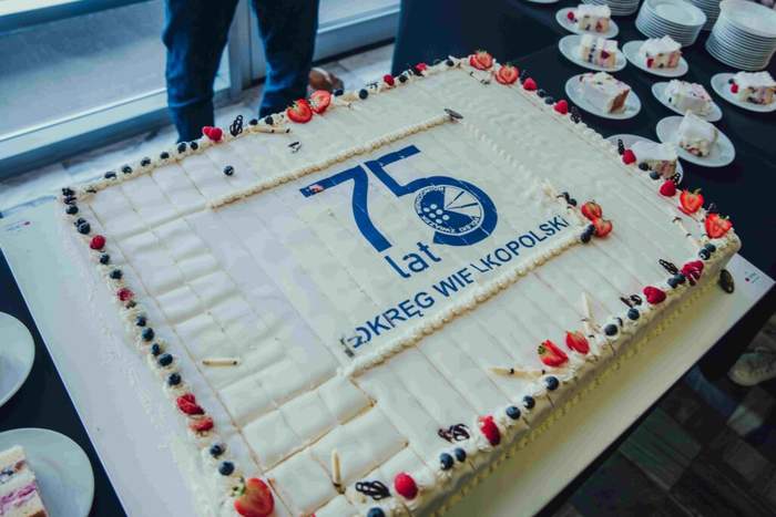 Pokaż zdjęcie: Duży prostokątny tort z jasnym kremem, udekorowany na brzegach owocami i rozetkami. Na środku znajduje się niebieski napis „75 lat” oraz okrągłe logo z podpisem „OKRĘG WIELKOPOLSKI”. W tle widać talerzyki z pokrojonymi kawałkami ciasta oraz stojące obok osoby.