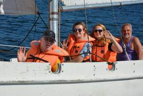 Pokaż zdjęcie: Cztery osoby na żaglówce, uśmiechają się i machają. Pan i dwie panie siedzą w pomarańczowych kapokach, za nimi dojrzały pan bez kapoku w fioletowej koszulce. 