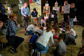 Pokaż zdjęcie: Artyści - dwóch mężczyzn i jedna dziewczynka malują stopami. Siedzą na tle obrazów. Uczestnicy spotkania obserwują artystów i robią im zdjęcia.