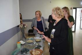 Pokaż zdjęcie: Przy ścianie na stole stoją prace ceramiczne, wokół stołu stoją cztery kobiety i oglądają prace