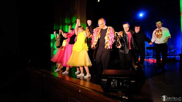 Grupa osób, kobiety i mężczyźni, w kolorowych strojach, stoją na scenie