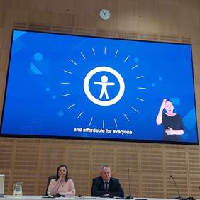 Pokaż zdjęcie: przy długim stole konferencyjnym siedzą dwie osoby. Kobieta prof. María Soledad Cisternas Reyes prowadząca wykład) w różowym golfie i czarnych włosach do ramion oraz mężczyzna Pan Paweł Wdówik – organizator wydarzenia. Ubrany w czarny garnitur, białą koszulę i krawat. Za nimi na ścianie wyświetlona niebieska plansza ze znakiem dostępności oraz napisem w j. angielskim: Affordable for everyone