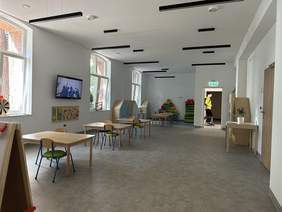 Pokaż zdjęcie: Przestronne jasne pomieszczenie w rzędzie stoją ławki z krzesłami, z boku na ścianie wisi duży telewizor, z sufitu zwisają ciemne szerokie lampy.