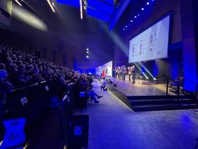 Pokaż zdjęcie: Na scenie stoi mężczyzna ubrany w garnitur. Trzyma list oprawiony w twardą okładkę i mówi do mikrofonu. Obok stoi kobieta w blond włosach i w czerwonej sukni, i mężczyzna ubrany w garnitur białą koszulę i muchę. Na telebimie wiszącym na scen