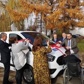 Pokaż zdjęcie: Uczestnicy WTZ oraz zaproszeni goście przecinają czerwona wstęgę związaną na białym samochodzie.