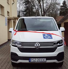 Pokaż zdjęcie: Na chodniku zaparkowany jest biały bus z naklejką informującą o przystosowaniu pojazdu do przewozu osób z niepełnosprawnością. Na masce samochodu widoczna jest biało-czerwona wstęga wraz z kokardą. W tle możemy dostrzec dwa budynki, garaż oraz drzewa iglaste i liściaste pozbawione liści.