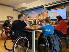 Pokaż zdjęcie: Pięć osób poruszających się na wózku znajduje się w kuchni przy stoliku i rozmawia z kobietą, która trzyma w dłoni filiżankę. W tle na monitorze wyświetlona jest prezentacja zatytułowana: „Porozmawiajmy o jedzeniu”.