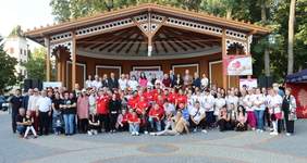 Pokaż zdjęcie: Grupa kilkudziesięciu osób stoi przed amfiteatrem. W tle drzewa liściaste, nagłośnienie oraz namiot jednego z wystawców.