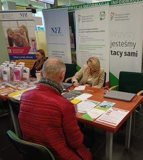 Pokaż zdjęcie: Kobieta reprezentująca Oddział Wielkopolski PFRON udziela informacji starszemu mężczyźnie z niepełnosprawnością narządu słuchu, pisząc na kartce odpowiedź. Na stoliku Oddziału ułożone są różne ulotki, w tle znajdują się dwa banery: PFRON oraz CIDON.