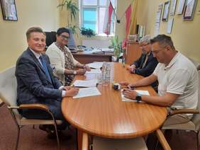 Pokaż zdjęcie: Przy owalnym stole siedzą cztery osoby: kobieta i trzech mężczyzn. Podpisują umowę. Na stole leżą różne dokumenty, pieczątki, woda mineralna. 