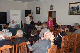 Pokaż zdjęcie: Kilka osób siedzi na krzesłach, przed nimi mężczyzna z mikrofonem oraz kobieta, za nimi baner z logo PFRON