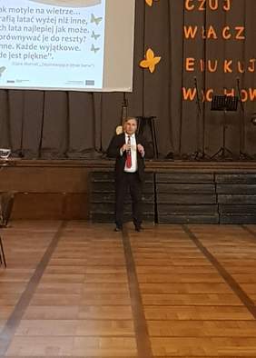 Pokaż zdjęcie: Meżczyzna w ciemnym garniturze stoi przed sceną trzymająć w ręku mikrofon.