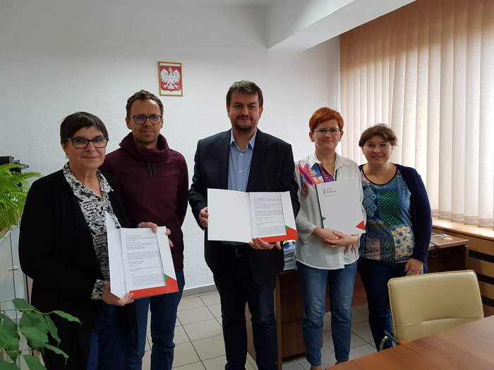 Pokaż zdjęcie: W Gabinecie Dyrektora na tle ściany obok biurka stoją osoby podpisujące umowę, Dyrektor trzyma w ręku umowy