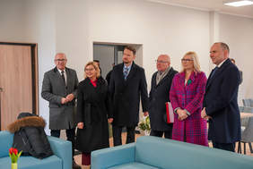 Sześć osób w zimowych płaszczach stoi w jasnym, nowoczesnym pomieszczeniu z turkusowymi kanapami i szarymi krzesłami w tle. Dwie kobiety i czterech mężczyzn, patrzą w kierunku widza. Na pierwszym planie widać turkusową kanapę, a na niej złożoną czarną kurtkę z futrzanym kapturem obok wazonu z czerwonymi tulipanami.