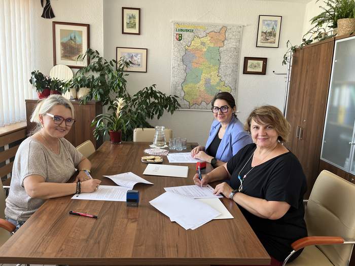 Pokaż zdjęcie: W pomieszczeniu Oddziału, przy stoliku, znajdują się trzy kobiety, które są uśmiechnięte i ubrane kolorowo oraz trzymają w ręku długopisy