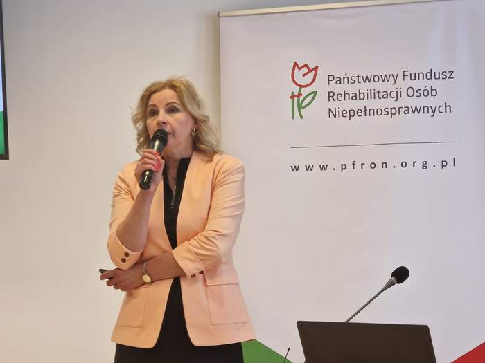 Pokaż zdjęcie: Zbliżenie na kobietę prowadzącą wykład, blondynka w łososiowej marynarce, trzyma w ręku mikrofon.