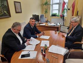 Pokaż zdjęcie: Przy owalnym stole siedzi trzech mężczyzn. Podpisują umowy. Na stole leżą różne dokumenty, pieczątki, długopisy i telefony. 