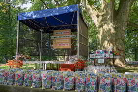 Na tle zielonych drzew widzimy scenę z napisem dzień osób niepełnosprawnych w gminie Zarzecze, po prawej stronie bardzo gruby pień drzewa. Pod sceną ustawione są liczne reklamówki z motywem regionalnym