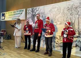 Pokaż zdjęcie: Grupa uczestników w świątecznych swetrach i czapkach mikołajowych występuje na scenie, trzymając mikrofony i kartki z tekstami kolęd; obok stoi prowadząca wydarzenie.