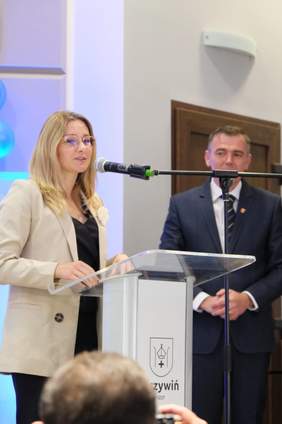 Pokaż zdjęcie: Wystąpienie pracownika Państwowego Funduszu Rehabilitacji Osób Niepełnosprawnych, a po prawej stronie stoi mężczyzna w garniturze. Kobieta przemawia przy przezroczystej mównicy z herbem Krzywina.