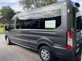 Pokaż zdjęcie: Grafitowy mikrobus z dużą naklejką z boku, z napisem dofinansowane ze środków PFRON. Wypolerowana karoseria pięknie odbija światło.