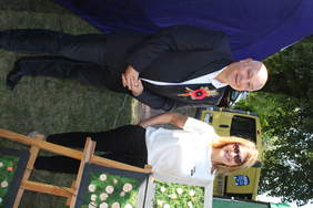 Pokaż zdjęcie: Zdjęcie mężczyzny w garniturze i kobiety w spodniach, blondynki w okularach pozują i uśmiechają się do zdjęcia. Osoby są na dożynkach i stoją między namiotami