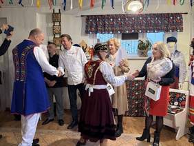 Pokaż zdjęcie: Osoby w strojach ludowych wymieniają uściski dłoni i prezenty z uczestnikami, w tym kucharzem. W tle dekoracje z kwiatowym motywem oraz kolorowe wstążki, nadające wydarzeniu folklorystyczny charakter.