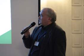 Pokaż zdjęcie: Mężczyzna w garniturze stoi trzymając w ręku mikrofon, spogląda w kierunku prezentacji wyświetlanej z tyłu za nim