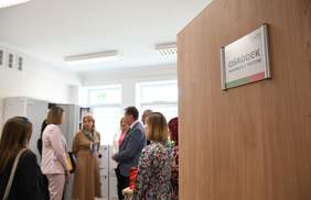 Pokaż zdjęcie: Przez otwarte drzwi na których jest tabliczka ośrodek wsparcia i testów widać wnętrze pomieszczenia i rozmawiającą grupę ośmiu osób